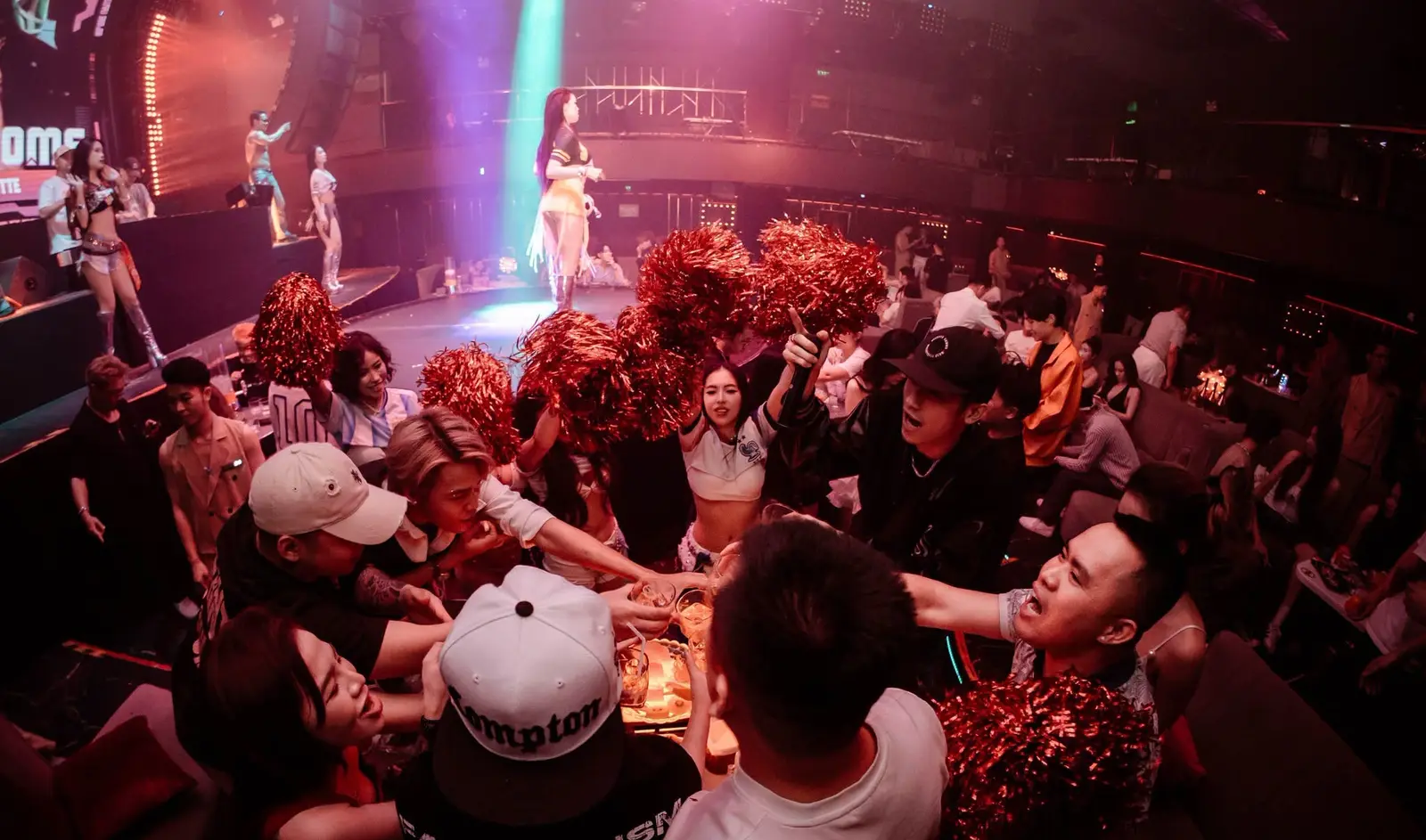 Khách và vũ đoàn cầm pom-pom đỏ giữa khu bàn đông người tại Canalis Club, sân khấu phía xa